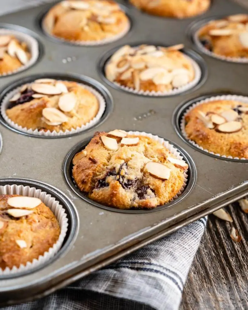 Close-up 95-degree angle of a tray of baked Cherry Almond muffins dotted with baked cherries and slivers of almonds over a blue napkin and wood board.