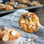Front facing photo of a baked cherry almond muffin sitting on it side showing the top of the muffin sprinkled with sliced almonds on a blue stripped napkin. A blurred muffin is in the front to the side and additional almonds are sprinkled over the napkin