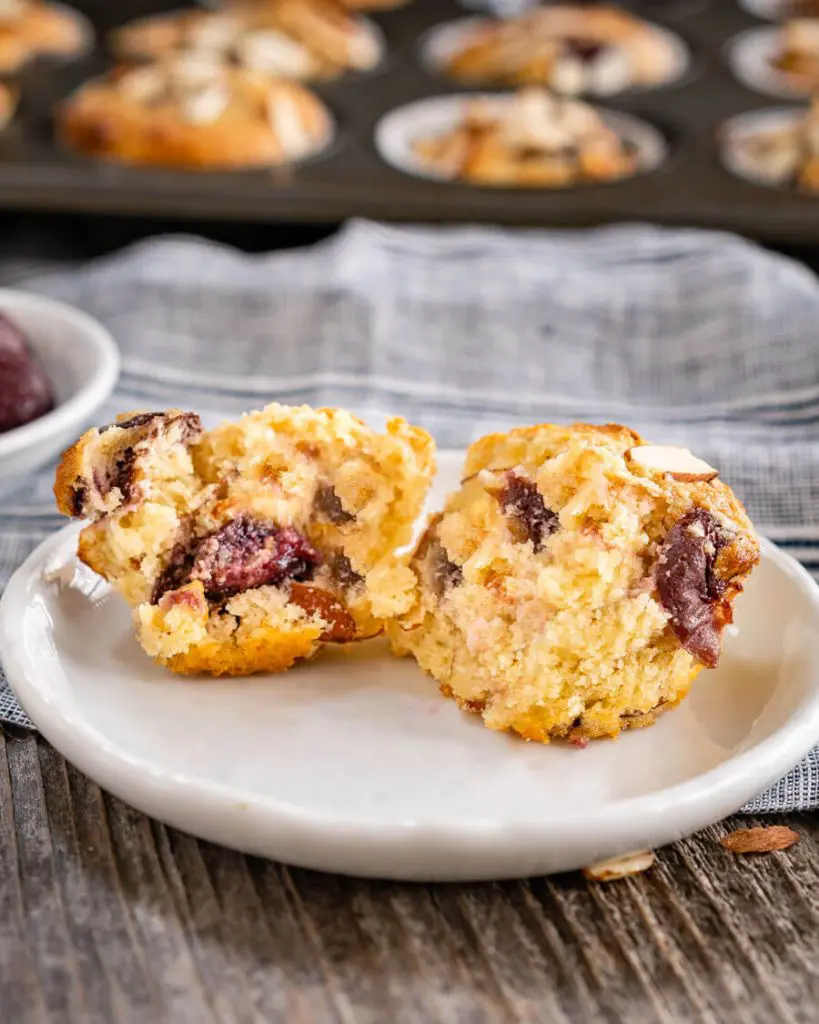 A small white plate with a cherry muffin cut in half showing the baked cherries and sliced almonds over a blue napkin. A tray of muffins is blurred in the background.