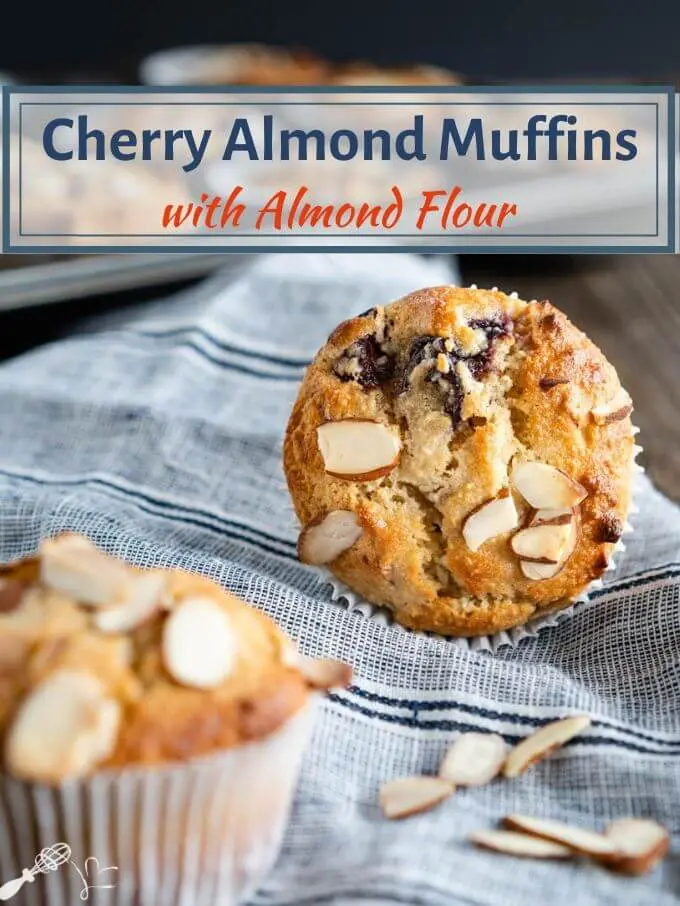 A close-up front view of a Cherry Almond Muffin laying on its site showing the top which has cherries and sliced almonds baked into it over a blue striped napkin. The title of the recipe \"Cherry Almond Muffins with Almond Flour\" is printed over the top of the photo.