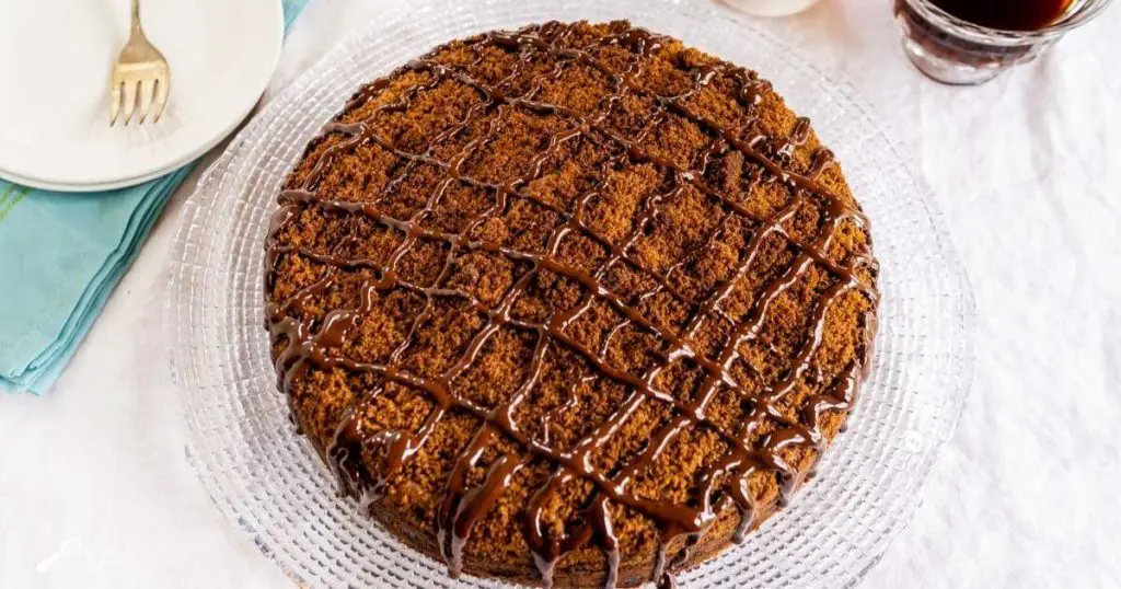 Top down view of a chocolate coffee cake topped with cinnamon streusel and drizzled with dark chocolate sitting on a glass plate over a white table cloth. A pretty turquoise napkin and two white plates with a fork sit to one side and a cup of coffee sits on the other.
