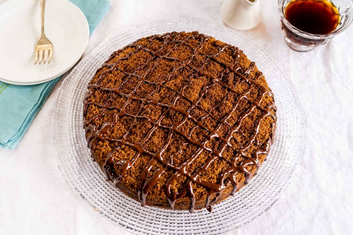 Top down view of a chocolate coffee cake topped with cinnamon streusel and drizzled with dark chocolate sitting on a glass plate over a white table cloth. A pretty turquoise napkin and two white plates with a fork sit to one side and a cup of coffee and creamer pitcher sits on the other.