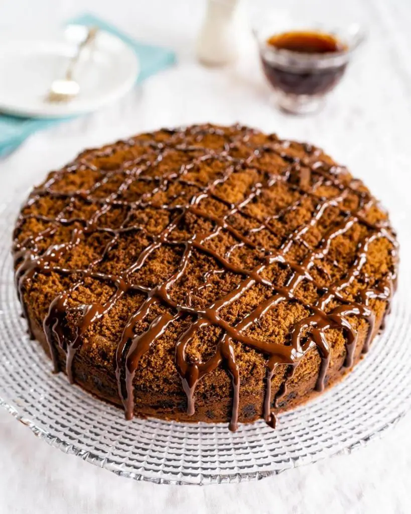 Top angle view of a chocolate coffee cake topped with cinnamon streusel and drizzled with dark chocolate sitting on a glass plate over a white table cloth. A pretty turquoise napkin and s white plate with a fork sit to one side and a cup of coffee and creamer pitcher sits on the other.