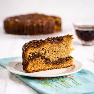 A side angle of a slice of Chocolate Coffee Cake on a white plate showing a layer of chocolate running through it and topped with streusel then drizzled with more chocolate. A pretty spring blue napkin sits below the plate and the cake sits behind it with a cup of coffee to the side.