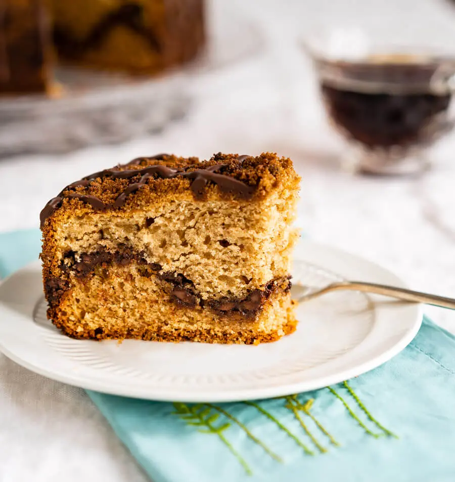 A side angle of a slice of Chocolate Coffee Cake with a bite taken out on a white plate with a fork sitting beside it showing a layer of chocolate running through it and topped with streusel then drizzled with more chocolate. A pretty spring blue napkin sits below the plate and the cake sits behind it with a cup of coffee to the side.