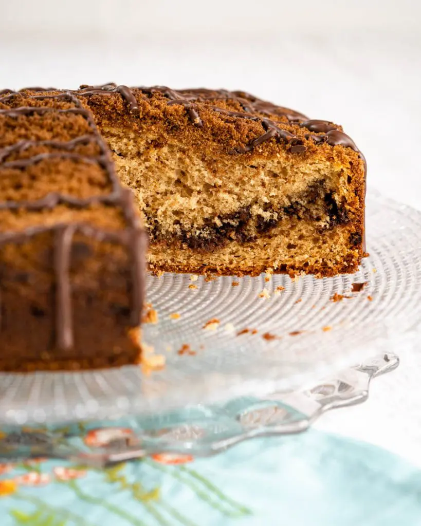 Side view of a Chocolate Coffee Cake with Sour Cream sitting on a glass cake plate. A piece has been removed showing the layer of dark chocolate that runs through it. The cake is topped with a cinnamon streusel and drizzled with more chocolate.