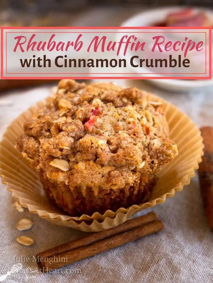 Front angle view of a rhubarb muffin topped with cinnamon crumble in a natural brown muffin paper. A cinnamon stick and oats sit in front.
