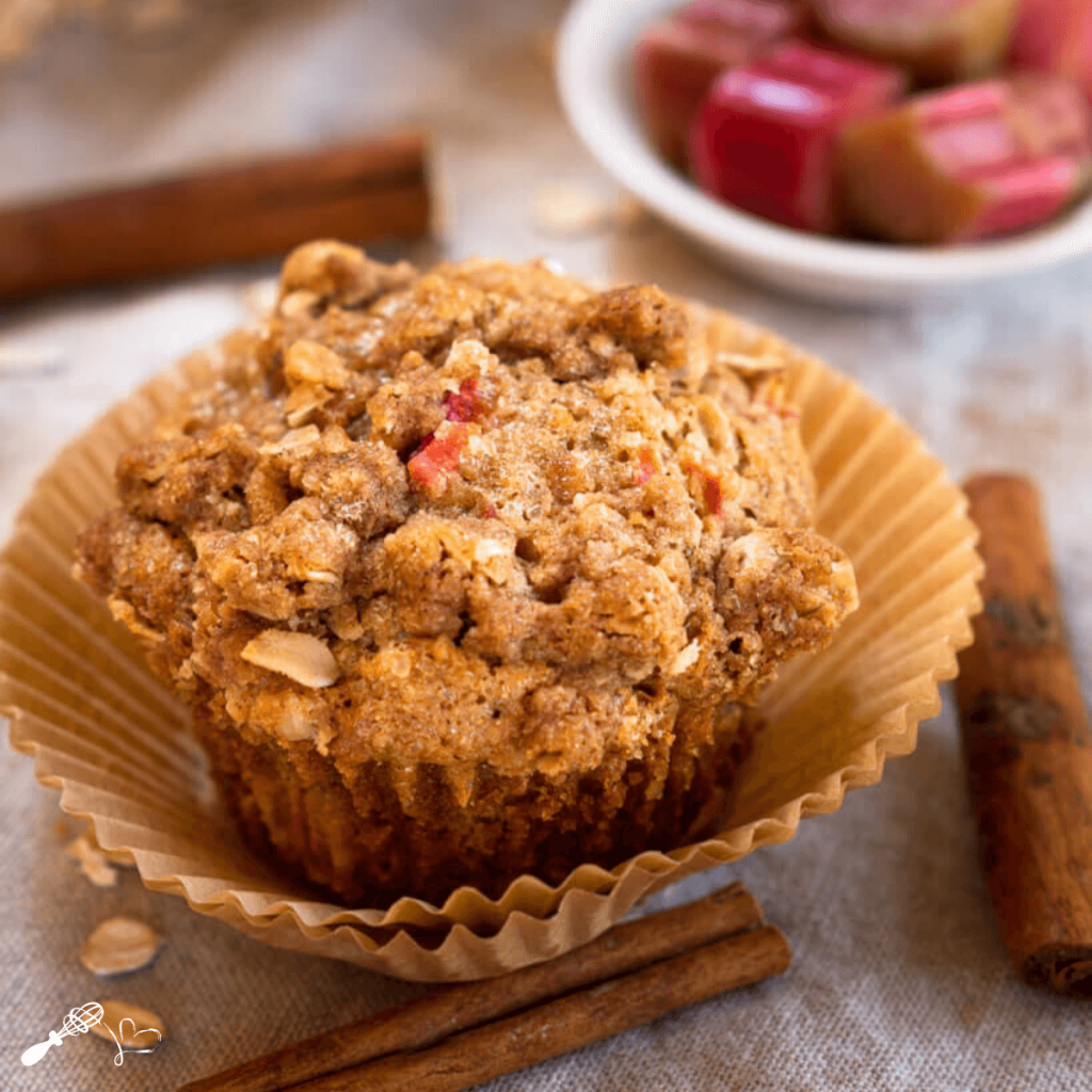 Front angle view of a rhubarb muffin topped with cinnamon crumble in a natural brown muffin paper. A cinnamon stick and oats sit in front.