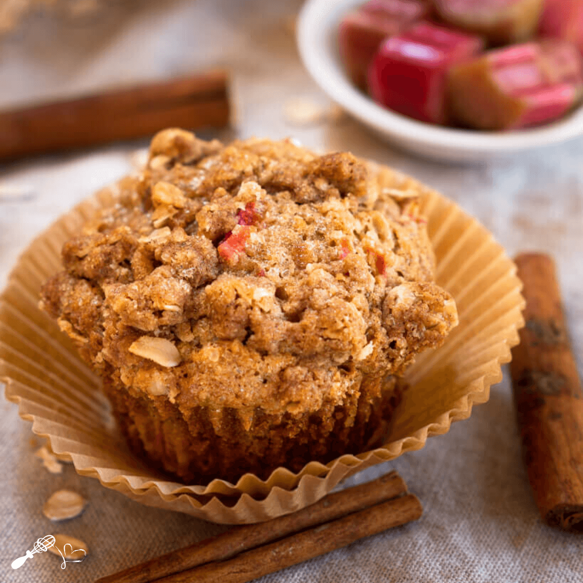 Front angle view of a rhubarb muffin topped with cinnamon crumble in a natural brown muffin paper. A cinnamon stick and oats sit in front.