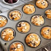 Side angle view of a muffin tin of baked cherry almond muffins topped with sliced almonds sitting on a striped blue napkin over a weathered wooden board. A white bowl of frozen cherries sit in the back.