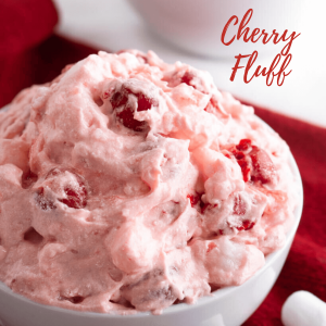 Top angle shot of a white bowl of pink cherry fluff on a red napkin with mini marshmallows scattered in front. The recipe title "Cherry Fluff" appears on the upper right side.