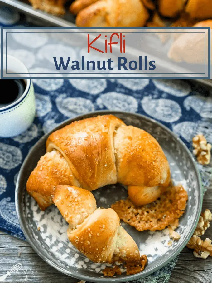 A top-down photo of a Kifli Walnut Roll sitting on a metal plate over a blue napkin. The title of the roll is on top and a cup of coffee sits to the back left.