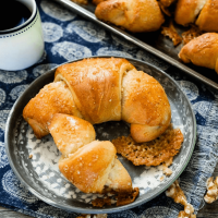 A metal bowl over a blue napkin with a large and small Kifli Walnut Roll sitting on it. A tray of rolls and a cup of coffee sit in the back.