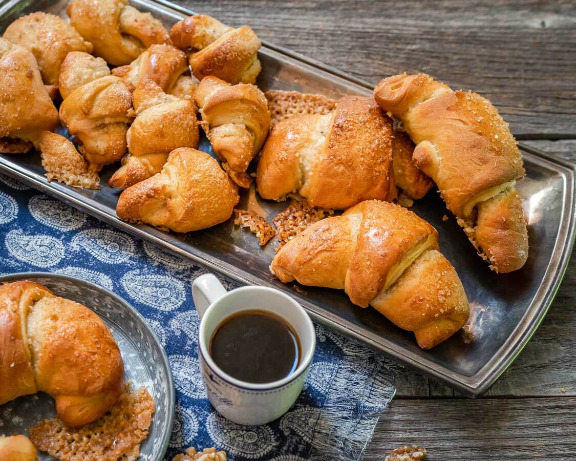 A silver tray of Kifli Walnut Rolls sitting on a blue napkin over a wooden board. A cup of coffee and a partial plate with a Kifli roll sit in the foreground.