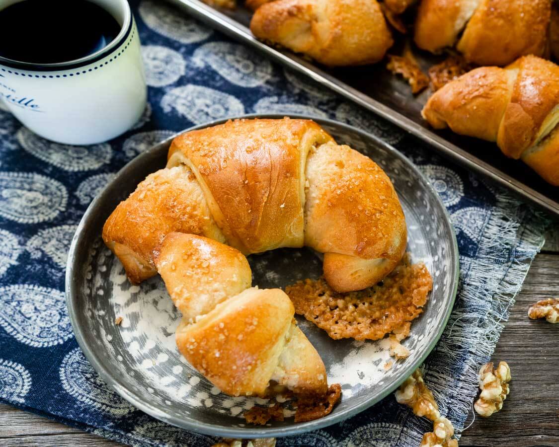 A metal plate holding a large and small Kifli Walnut roll sits on a blue paisley napkin. A tray of Kifli rolls and a cup of coffee sit in the back.
