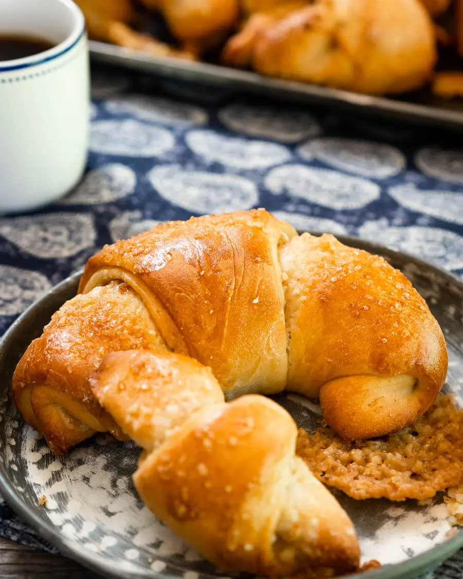 A close up of a large Kifli Walnut roll sitting on a metal plate in front of a tray of Kifli Walnut rolls over a blue paisley napkin.