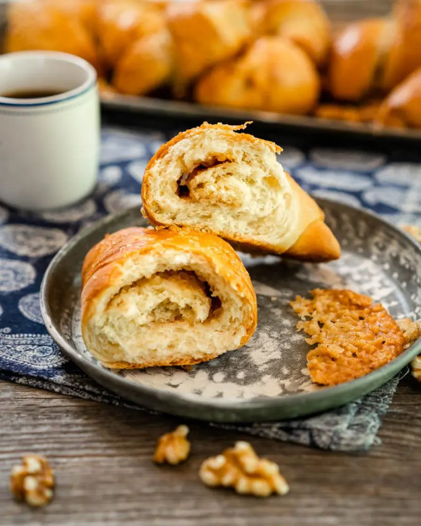 A Kifli Walnut Roll cut in half showing the cinnamon swirl inside. A cup of coffee and a tray of whole Kifli rolls sit in the background.