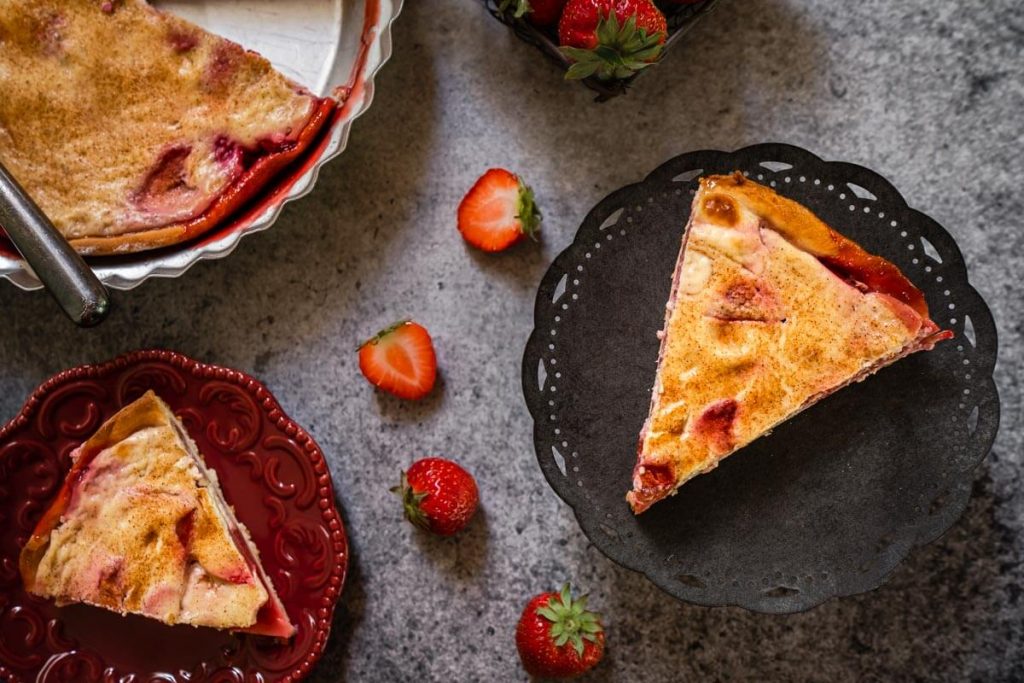 Top down view of slices of strawberry kuchen. One slice is on a cake stand and another slice is on a maroon plate. A corner shows a partial pie tin filled with kuchen. Strawberries are scattered about the photo.