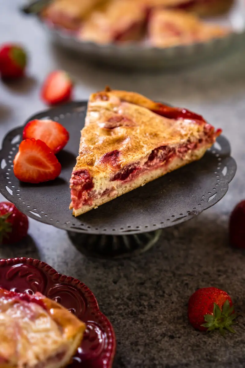 Side view of a slice of Kuchen showing a layer of strawberries over a crust and topped with custard sitting on a small gray cake stand. Strawberries are scattered around and a partial view of the whole kuchen in a metal pie tin sits in the background.