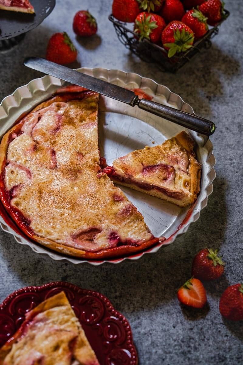 Top down view of a baked Kuchen with two slices missing. A knife sits over the back of the pie tin. Scattered strawberries and a basket of strawberries sit over a gray background. Partial slices of Kuchen appear on the edges of the photo