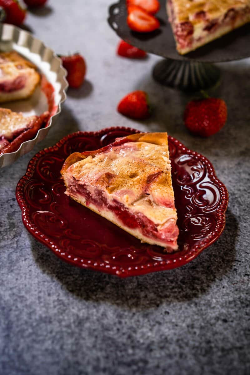 Angled view of a slice of Kuchen showing a layer of strawberries over a crust and topped with custard on a maroon plate. Strawberries are scattered around and a partial slice on a gray stand and the whole dessert in a pie tin is in the background on a gray background.