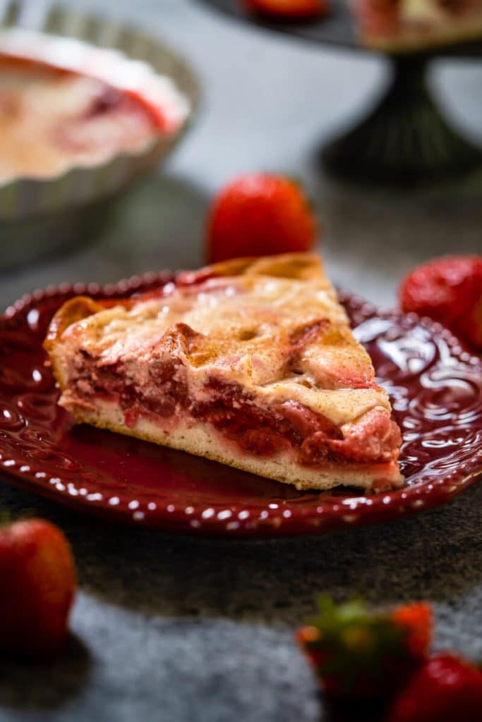 Side view of a slice of Kuchen showing a layer of strawberries over a crust and topped with custard on a maroon plate. Strawberries are scattered around the plate on a gray background.