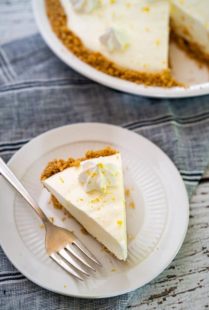 Slice of Lemon Icebox pie with a creamy lemon filling over a thick graham cracker crust on a white plate sitting over a blue striped napkin. A fork, and a partial second piece of pie sitting on a white plate is in the background.