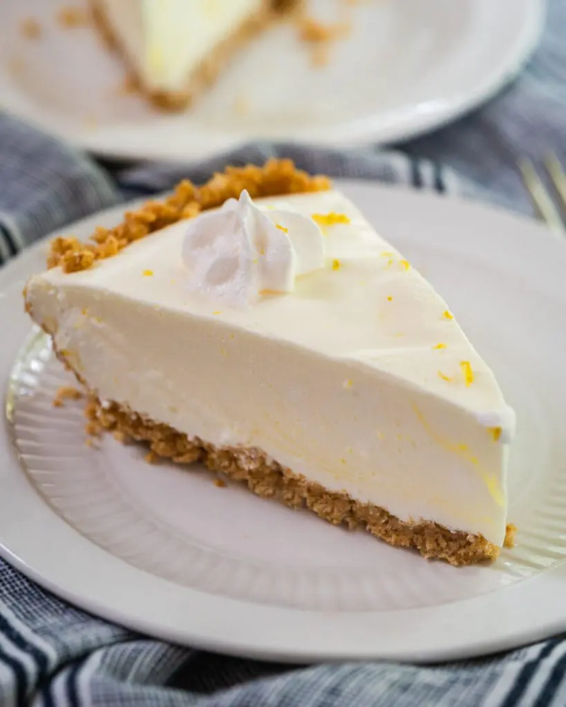 Close up side view of a slice of Lemon Icebox Pie. The slice has a thick graham cracker crust topped with a cool creamy layer and topped with a star of cool whip topping. The slice sits on a white plate over a blue napkin.