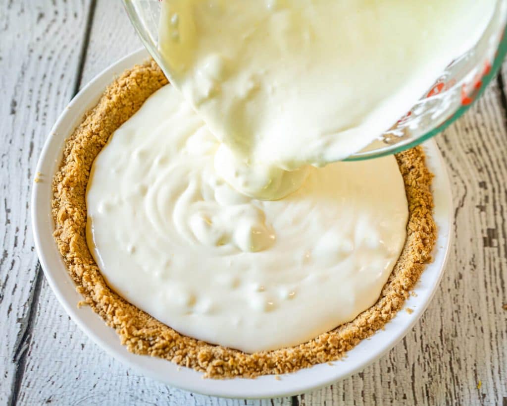 Top down photo of the creamy lemon pie filling being poured from a pyrex cup into a pie plate with a graham cracker crust. The pie pan sits over a white board background.