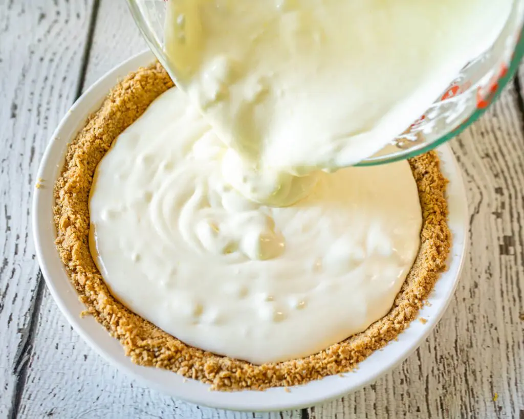 Top down photo of the creamy lemon pie filling being poured from a pyrex cup into a pie plate with a graham cracker crust. The pie pan sits over a white board background.