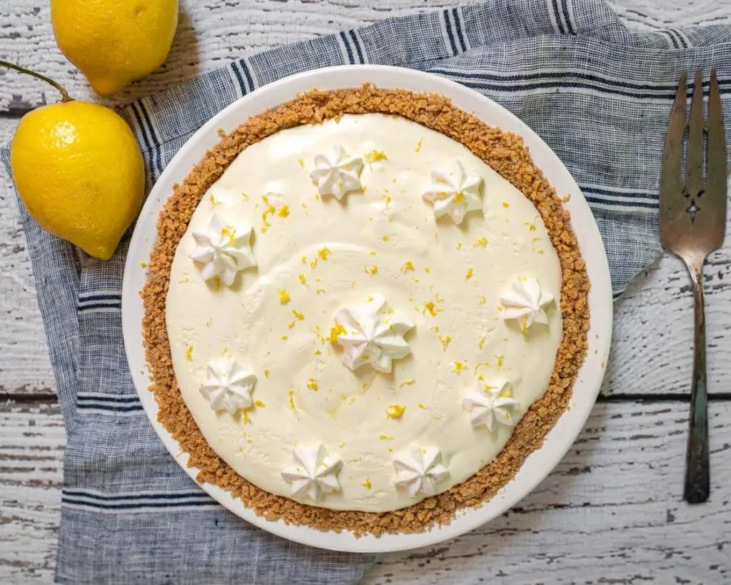 Top down view of a white pie plate filled with a yellow lemon Icebox Pie in a graham cracker crust. The pie has been decorated with stars of cool whip topping and grated lemon. The pie sits over a blue striped napkin and two lemons sits in the background and the pie sits next to an antique pie server.