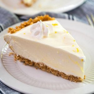 Close up side view of a slice of Lemon Icebox Pie. The slice has a thick graham cracker crust topped with a cool creamy layer and topped with a star of cool whip topping. The slice sits on a white plate over a blue napkin.
