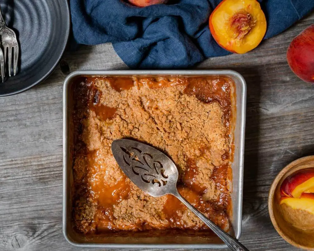 Top-down view of a pan of apple crisp with an antique pie server sitting on the top. Fresh peaches sit around the pan and a dark gray dish with forks sit in the background over a wooden background,