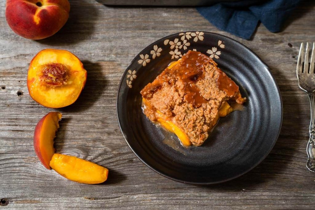 A top-down photo of a piece of peach crisp in a dark gray dish over a wooden background. Fresh peaches and a fork surround the dish. A blue napkin is in the background.