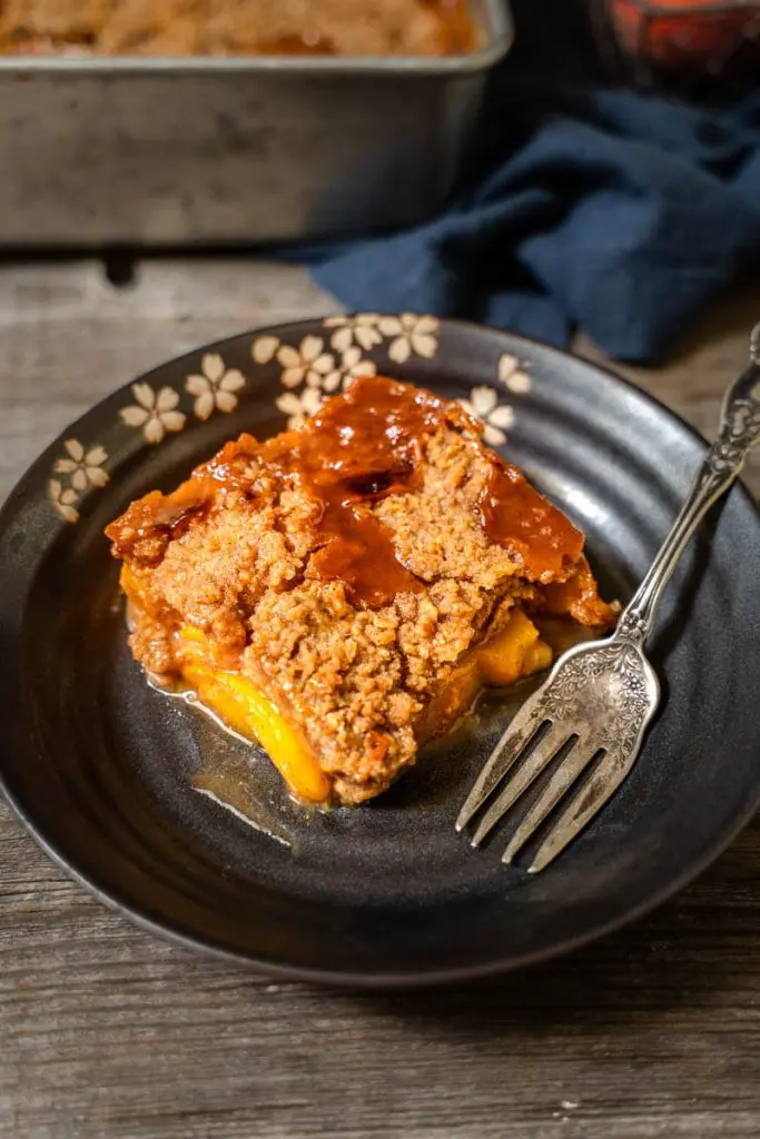 Top-down angle photo of a piece of peach crisp in a dark gray dish over a wooden background. A antique fork sits next to the crisp and a blue napkin is in the background.