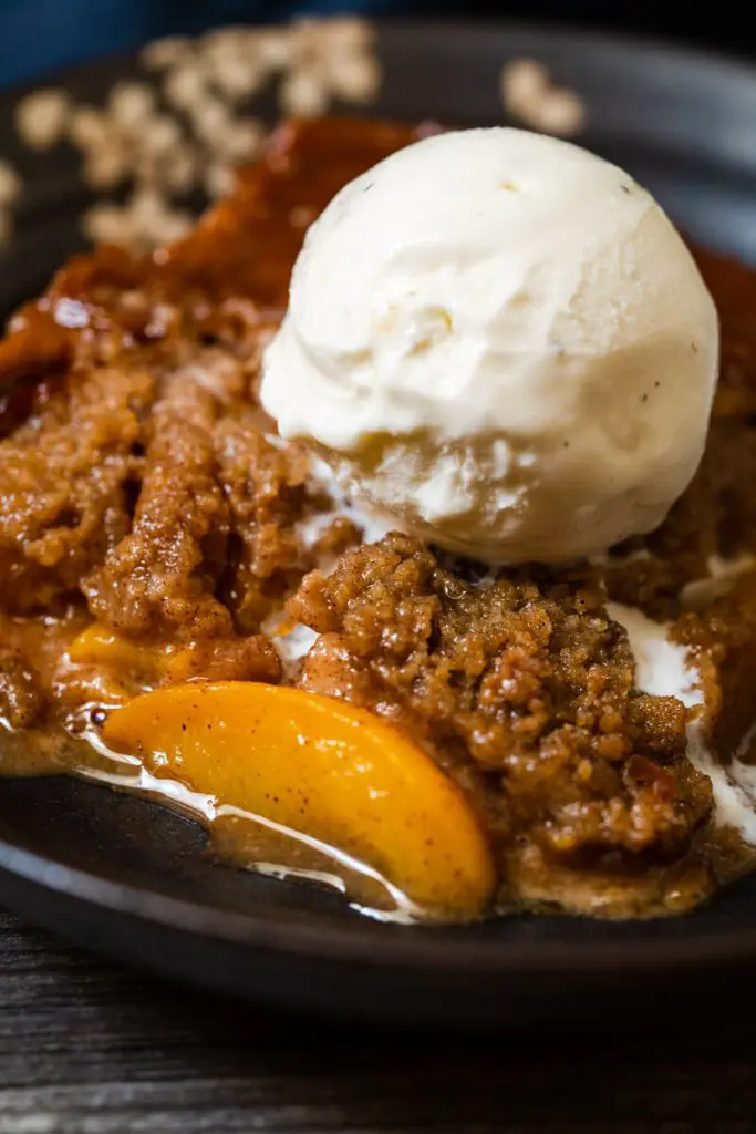 Top-down shot of a piece of peach crisp sitting in a dark gray bowl with vanilla ice cream melting down the side.