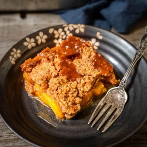 Top-down angle photo of a piece of peach crisp in a dark gray dish over a wooden background. A antique fork sits next to the crisp and a blue napkin is in the background.