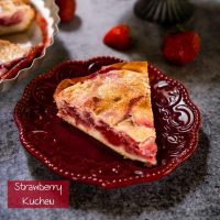 Angled view of a slice of Kuchen showing a layer of strawberries over a crust and topped with custard on a maroon plate. Strawberries are scattered around and a partial slice on a gray stand and the whole dessert in a pie tin is in the background on a gray background. The title 