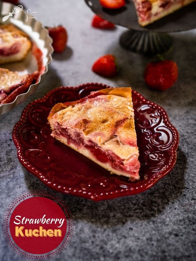 Angled view of a slice of Kuchen showing a layer of strawberries over a crust and topped with custard on a maroon plate. Strawberries are scattered around and a partial slice on a gray stand and the whole dessert in a pie tin is in the background on a gray background.