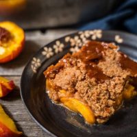 Top-down angle photo of a piece of peach crisp in a dark gray dish over a wooden background. A antique fork sits next to the crisp and a blue napkin is in the background.