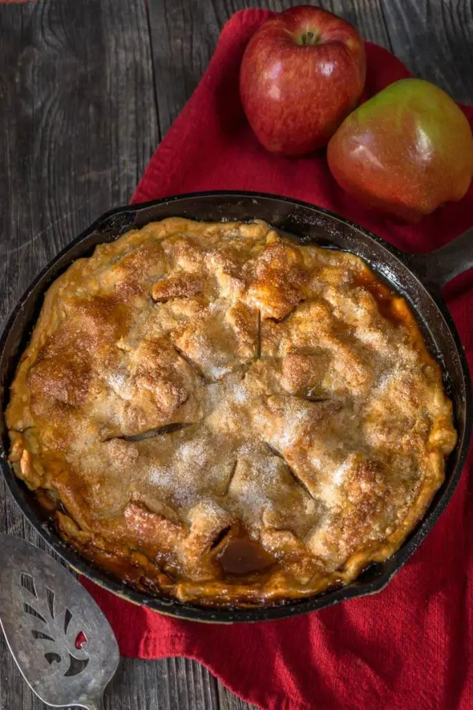 Top down view of an apple rhubarb pie baked in a cast-iron skillet sitting over a red napkin. Fresh apples are in the background and an antique pie server in the front.