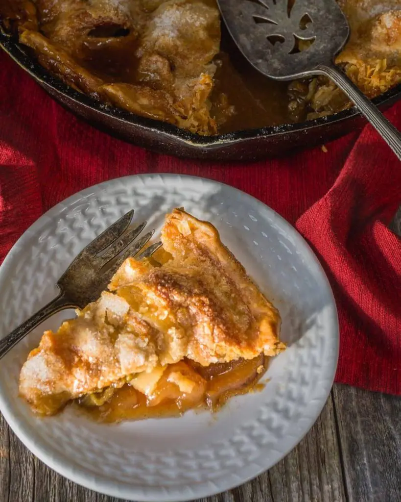 Top down photo of a slice of apple rhubarb pie on a white plate with an antique fork sitting next to it. A partial view of the whole pie in a cast-iron skillet is in the background sitting on a red napkin over a wooden background.