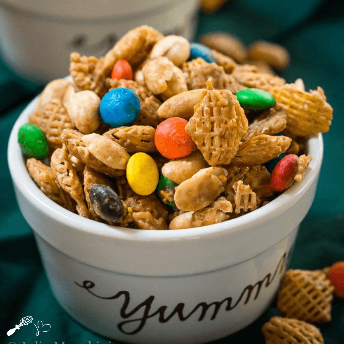 Close-up side view of a ramekin with the word "yummy" printed on the front filled with caramel crispix snack mix. A few pieces of the mix are scattered around the dish over a green napkin.