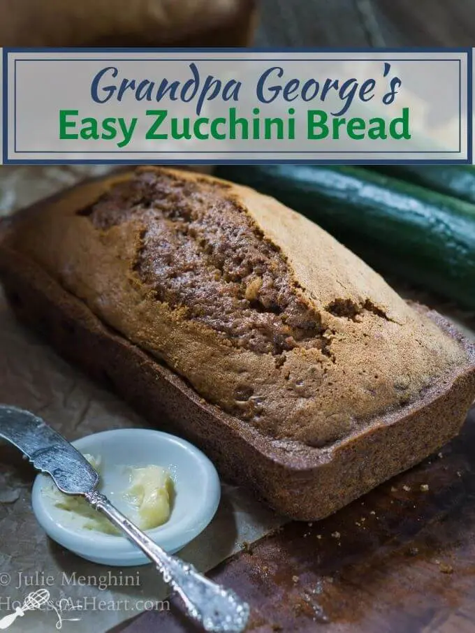 An angled loaf of bread with two fresh zucchini's sitting in the back. A white dish with butter and an antique knife sit in the front. The title "Grandpa George's Easy Zucchini Bread" runs across the top.