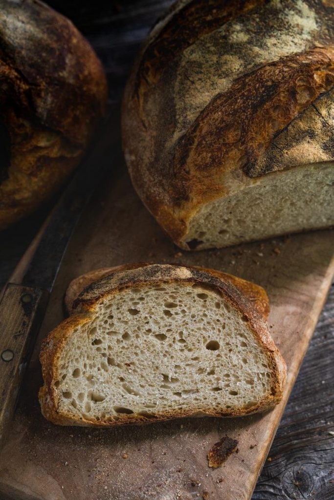 Top down photo of a slice of sourdough bread with the cut loaf sitting behind it on a wooden cutting board over a wooden background. A second loaf sits in the background next to an old knife.