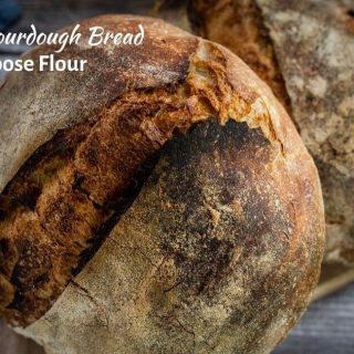 A top down photo of a round sourdough loaf leaning against an oval loaf of sourdough bread sitting on a wooden cutting board. An old knife and a blue napkin are sitting next to the cutting board on a wooden background. The title "Homemade Sourdough Bread with All-Purpose Flour is in the upper left corner.