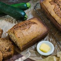 Top angle view of a loaf of zucchini bread sitting on a cooling rack next to a cut loaf on a wooden cutting board. Fresh zucchini sit in the background and a white dish of butter sits in the foreground next to an antique knife.