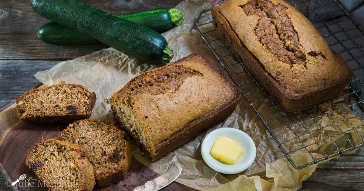 Top angle view of a loaf of zucchini bread sitting on a cooling rack next to a cut loaf on a wooden cutting board. Fresh zucchini sit in the background and a white dish of butter sits in the foreground next to an antique knife.