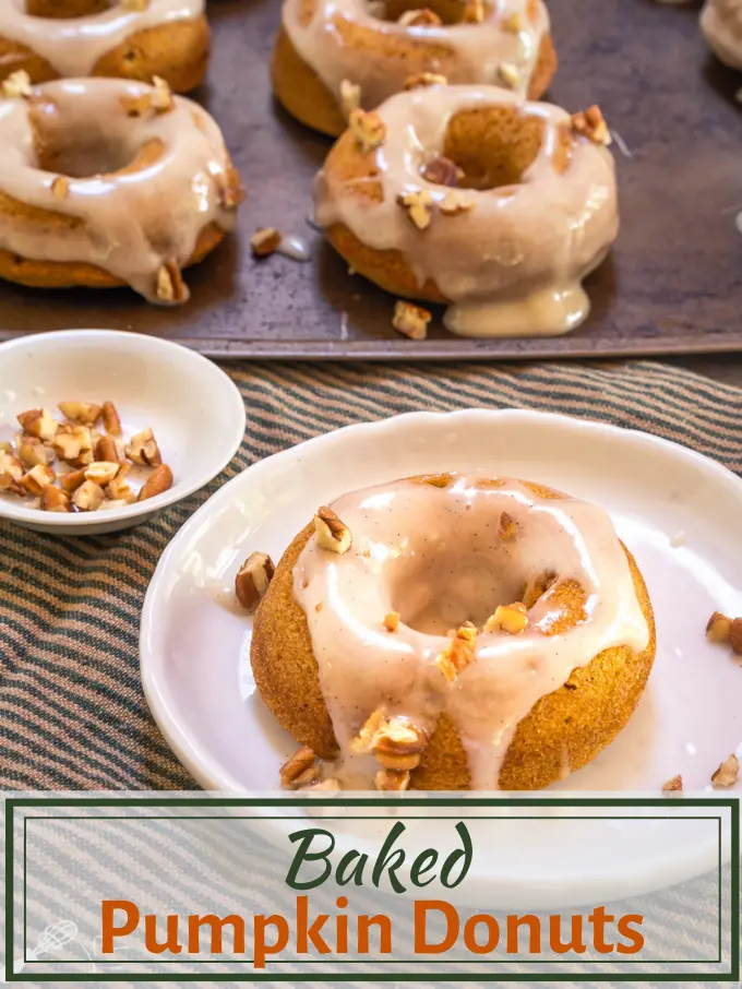 Top down photo of a pumpkin donut with cream cheese glaze and garnished with pecans on a white plate over a striped green napkin. A baking sheet of more donuts sit in the background.