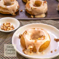 Top down photo of a pumpkin donut with cream cheese glaze and garnished with pecans on a white plate over a striped green napkin. A baking sheet of more donuts sit in the background.